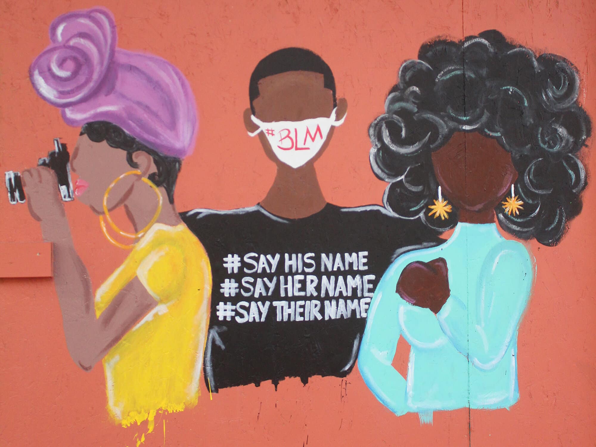 A photo shows a street mural depicting three Black women. The figure in the middle is wearing a face masks with "#BLM" printed on it and a T-shirt reading "#SAYHISNAME #SAYHERNAME #SAYTHEIRNAME." The figure on the left looks toward the side, holding a camera. The figure on the right appears to have her hand raised in a gesture of solidarity.