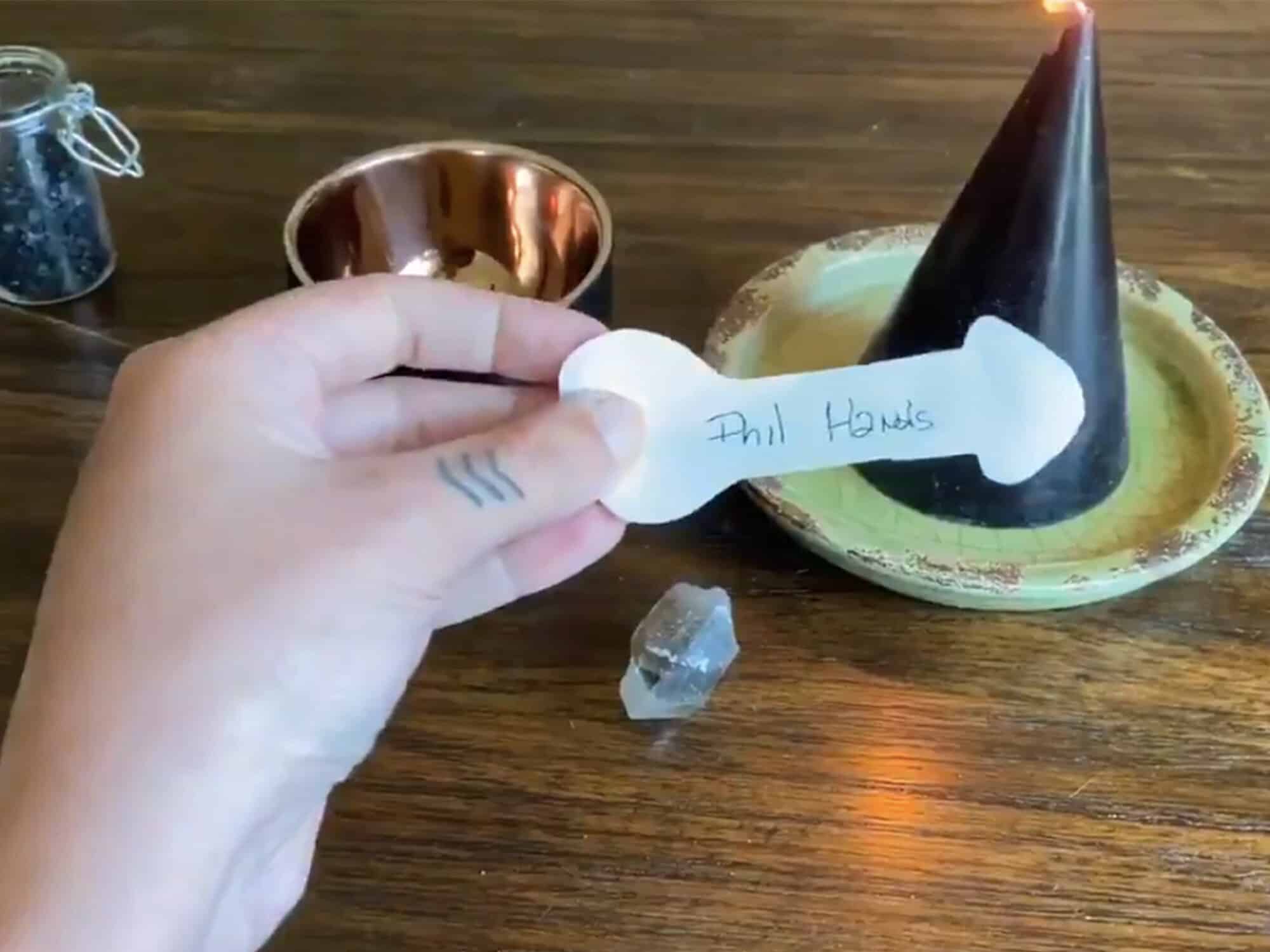 Someone's left hand holds a cutout piece of paper in the shape of a penis and testicles over a table with a small tray and conical vessel. The name "Phil Hands" is written on the shaft of the paper.