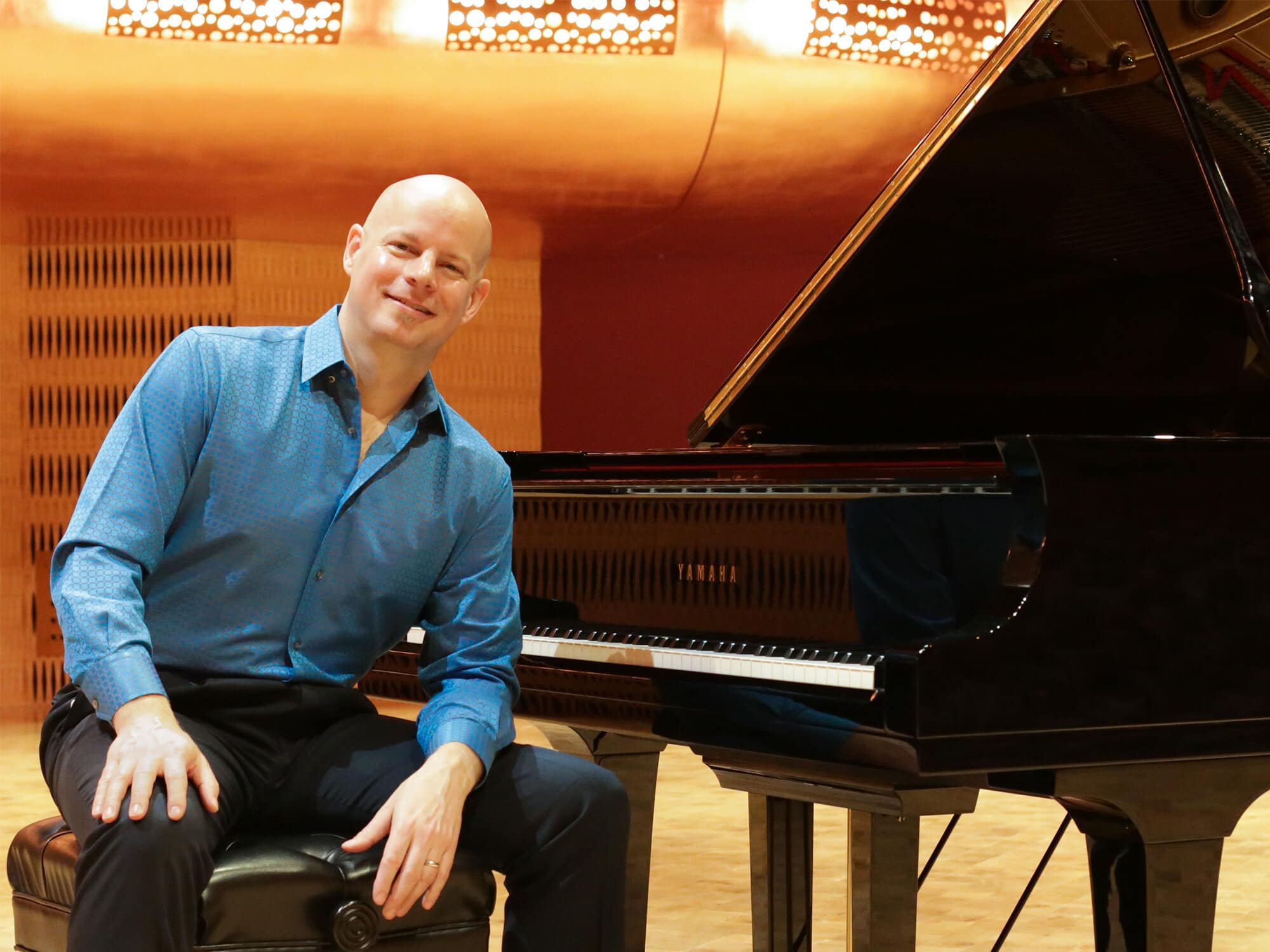 Johannes Wallmann is shown sitting at a grand piano, turned away from the keyboard to face the camera. He is wearing a blue dress shirt and the brightly lit interior of a concert hall is visible in the background.