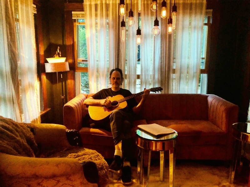 Aaron Scholz sits on a couch holding his acoustic guitar and staring at the camera. Brown, burgundy, and burnt orange hues bring the living room Scholz inhabits to life.