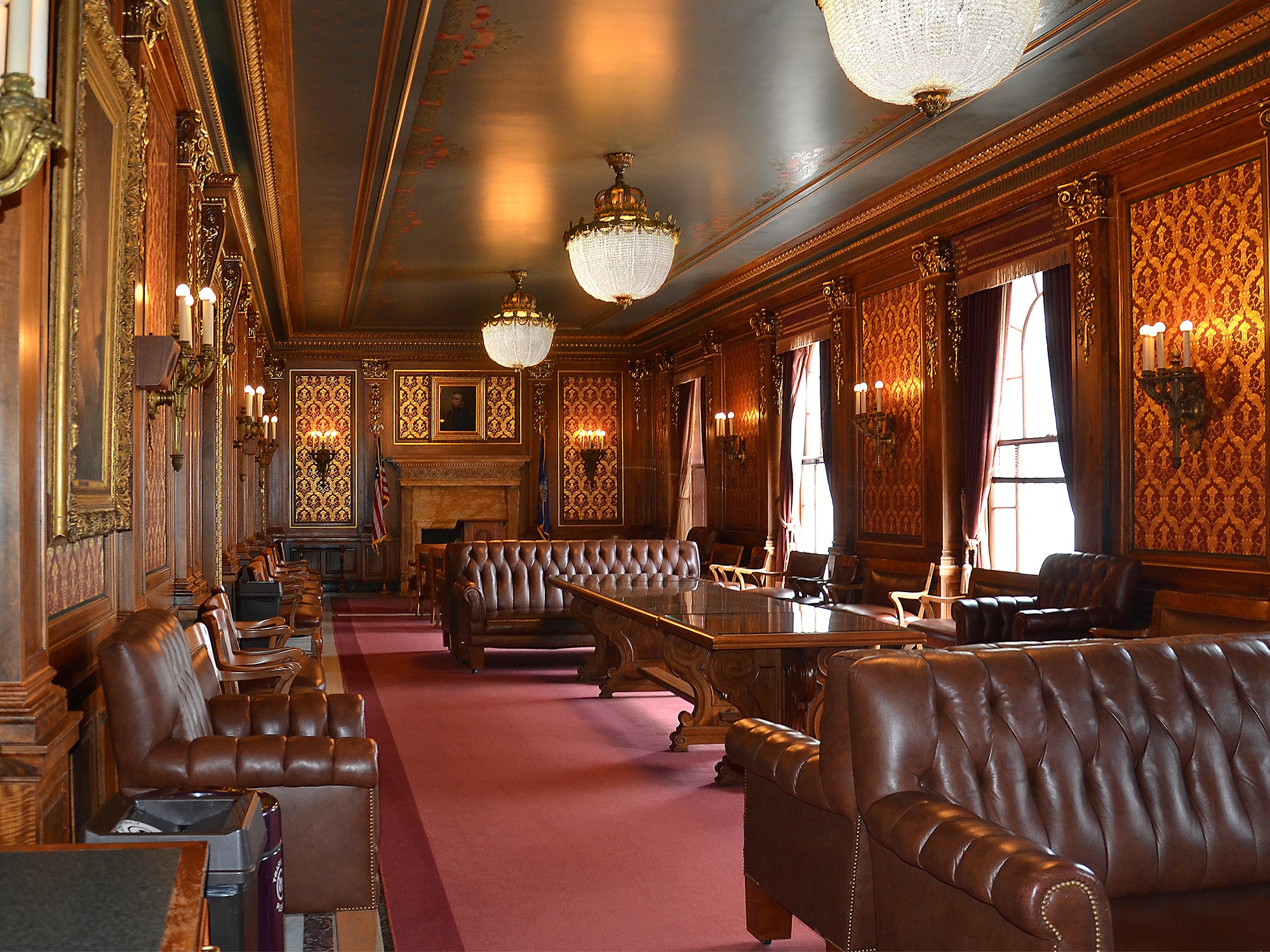 A photo shows a parlor in the Wisconsin State Capitol. The room has red carpet and a number of upholstered, expensive-looking couches, with ornate lighting and decorations on the walls. Photo by Richard Hurd on Flickr.