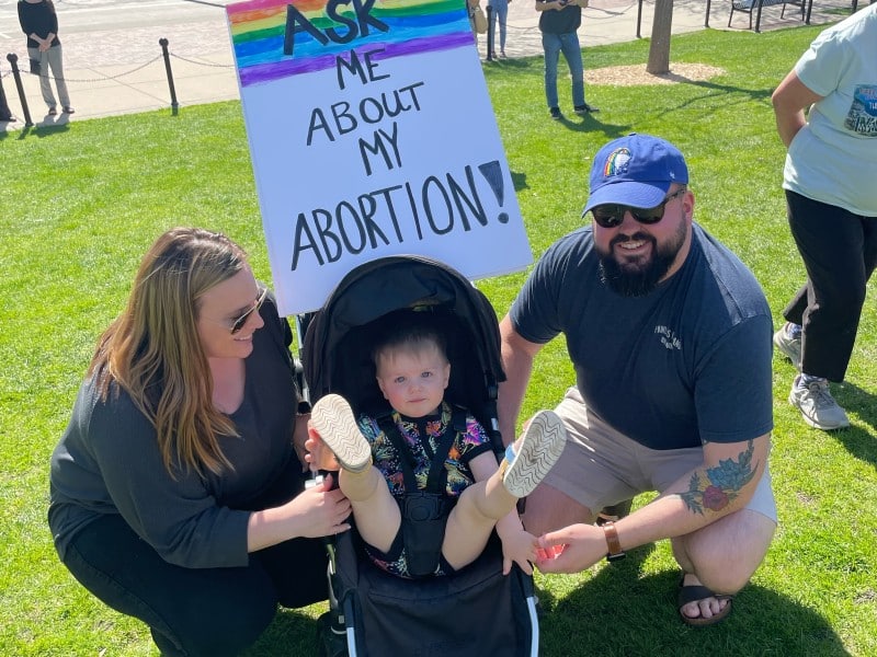 Two people crouch on either side of a stroller on the lawn of the Wisconsin State Capitol. The toddler in the stroller looks at the camera with their legs raised in the air. A sign above the stroller says "ASK ME ABOUT MY ABORTION!" with a rainbow at the top.