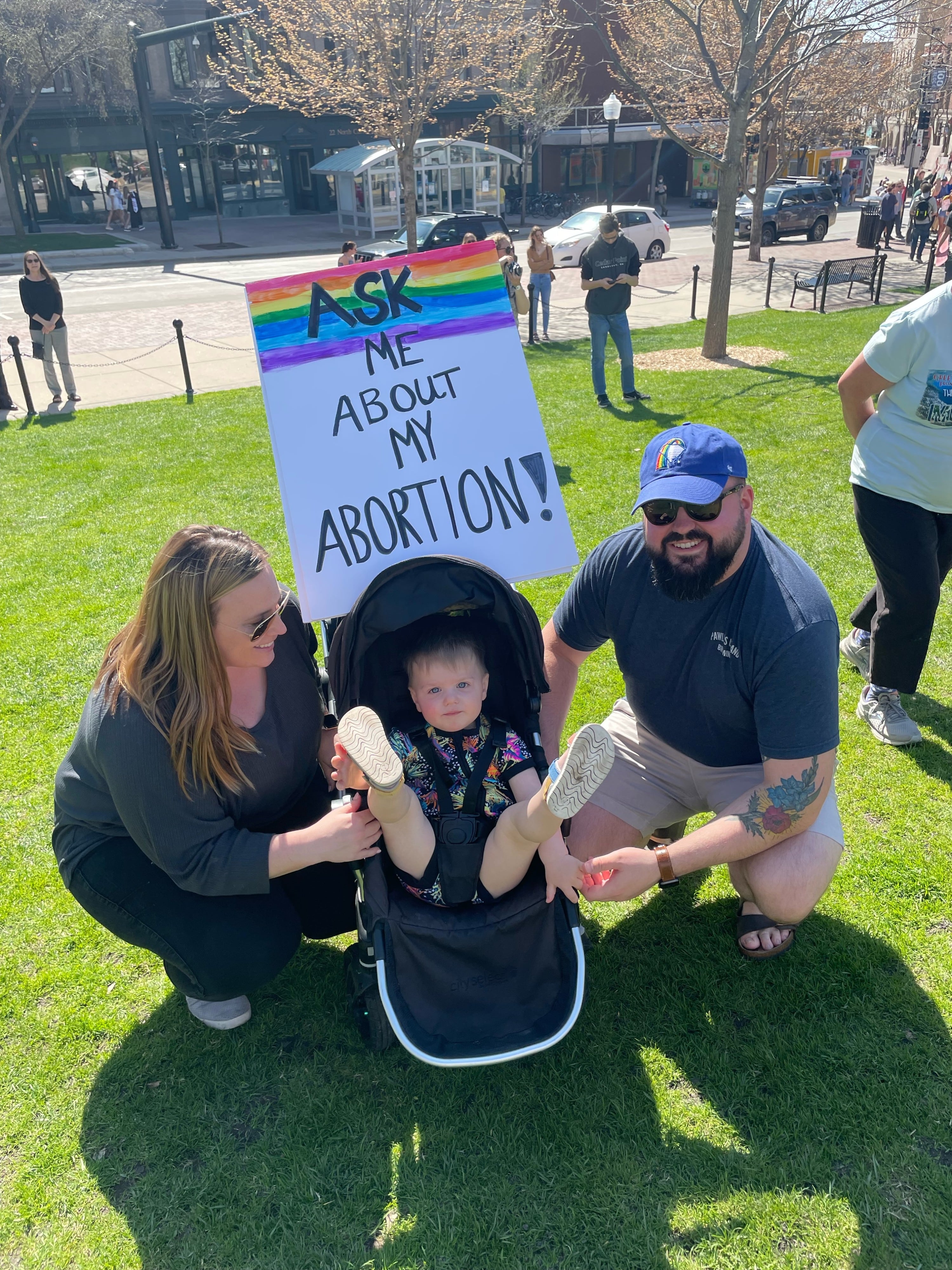 Two people crouch on either side of a stroller on the lawn of the Wisconsin State Capitol. The toddler in the stroller looks at the camera with their legs raised in the air. A sign above the stroller says "ASK ME ABOUT MY ABORTION!" with a rainbow at the top.