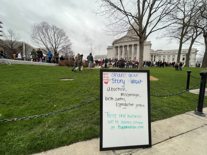 A whiteboard inviting oral histories / abortion stories is propped against metal fencing next to the lawn of the Wisconsin State Capitol Building. A protest in front of the Capitol is visible in the background.