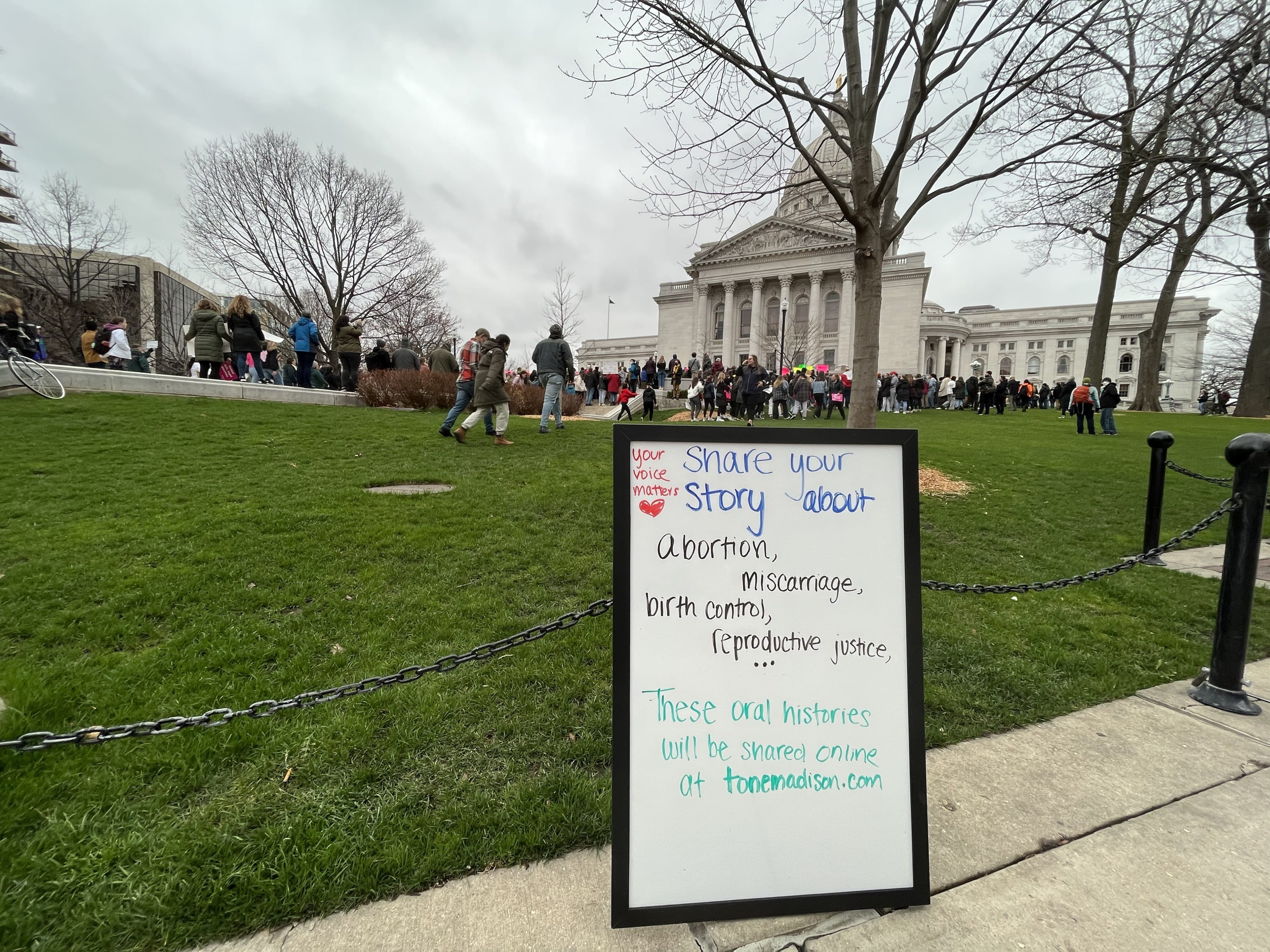A whiteboard inviting oral histories / abortion stories is propped against metal fencing next to the lawn of the Wisconsin State Capitol Building. A protest in front of the Capitol is visible in the background.