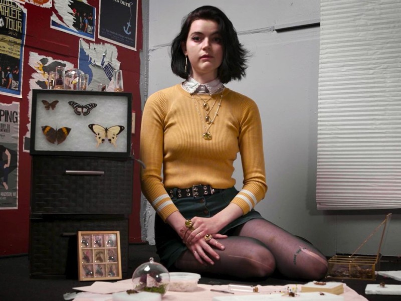 Sigra faces the camera in a still from the "Honeycore" music video directed by Jake Viaene. She poses in the corner of a room embellished with artifacts, metallurgy, and taxidermy (lepidoptera) from her own 27 Bones Studio amidst a red wall of partially torn show posters to her right and plain white blinds dangling below a windowsill to her left.