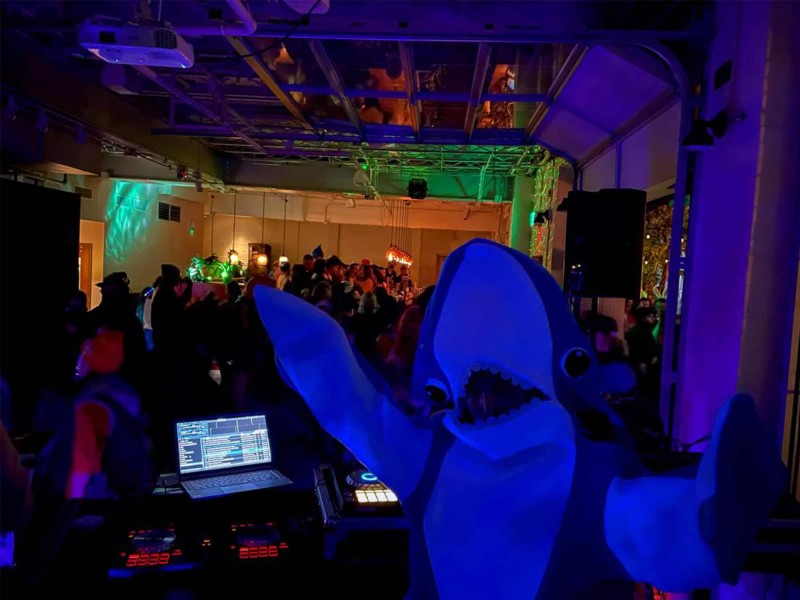 Madison-based DJ Sarah Akawa is shown posing in a shark costume. Behind Akawa are DJ controllers and a laptop, and in the background is a crowd filling a room at Robinia Courtyard