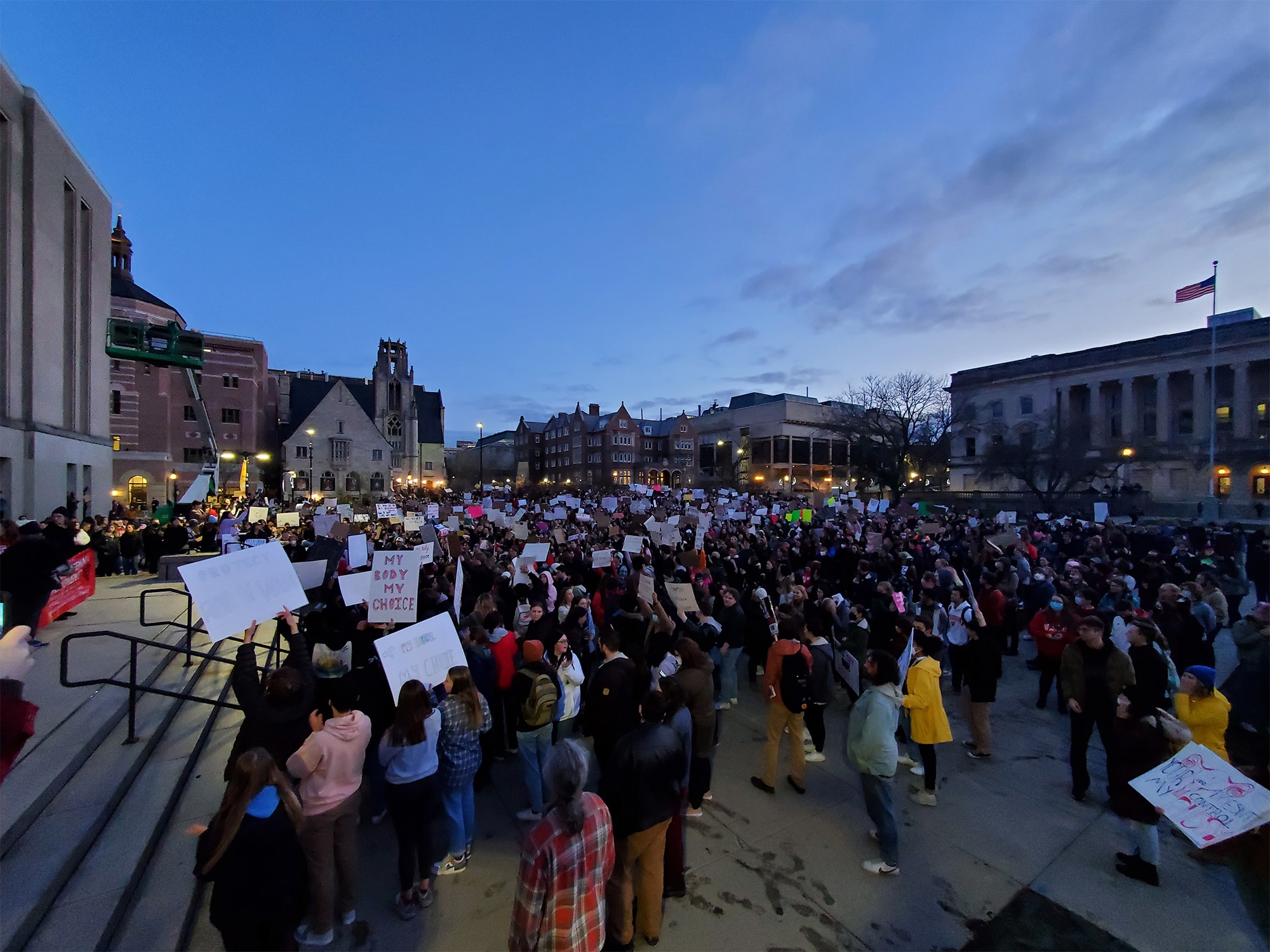 A photo shows a crowd of protestors during a May 3, 2022 rally for abortion rights on Library Mall in Madison. Handmade protest signs are visible in the foreground, including one that reads “MY BODY MY CHOICE.” In the background, it is twilight and several campus buildings are visible.