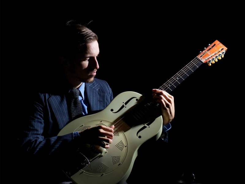 Musician William Z. Villain is shown seated in a dark blue suit, playing a white acoustic guitar and looking down at the fretboard.