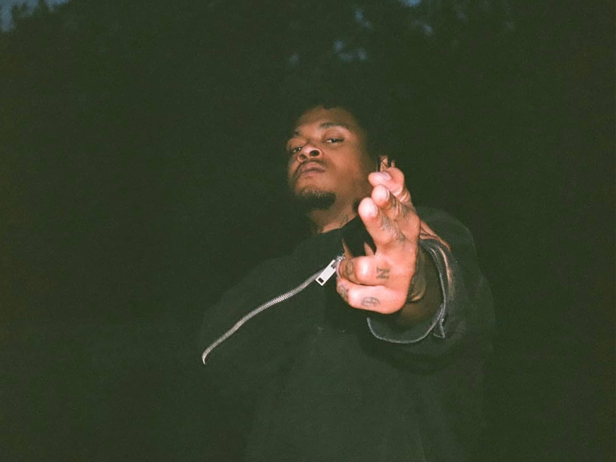 Nino Carter is photographed facing downward at the camera and pointing his hand at it. In the background it is late evening and a large tree can be seen in the darkness. Photo by @DHVNSEN.
