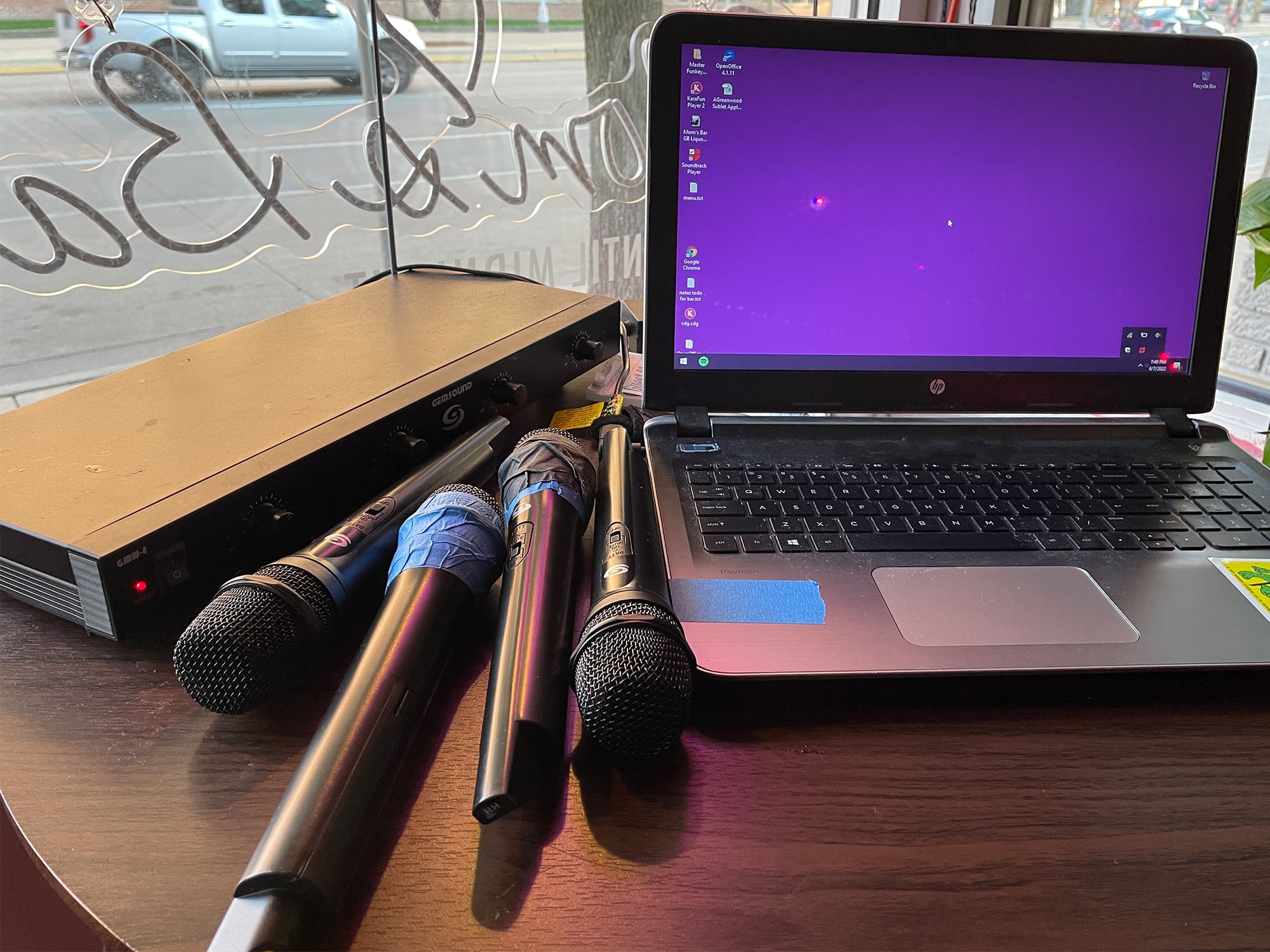 A laptop computer sits on a table at Mom's Bar. To its left, four microphones also rest on the table, next to a transmitter for wireless microphones. In the background, the neon sign for Mom's Bar is visible in the window from the reverse side.