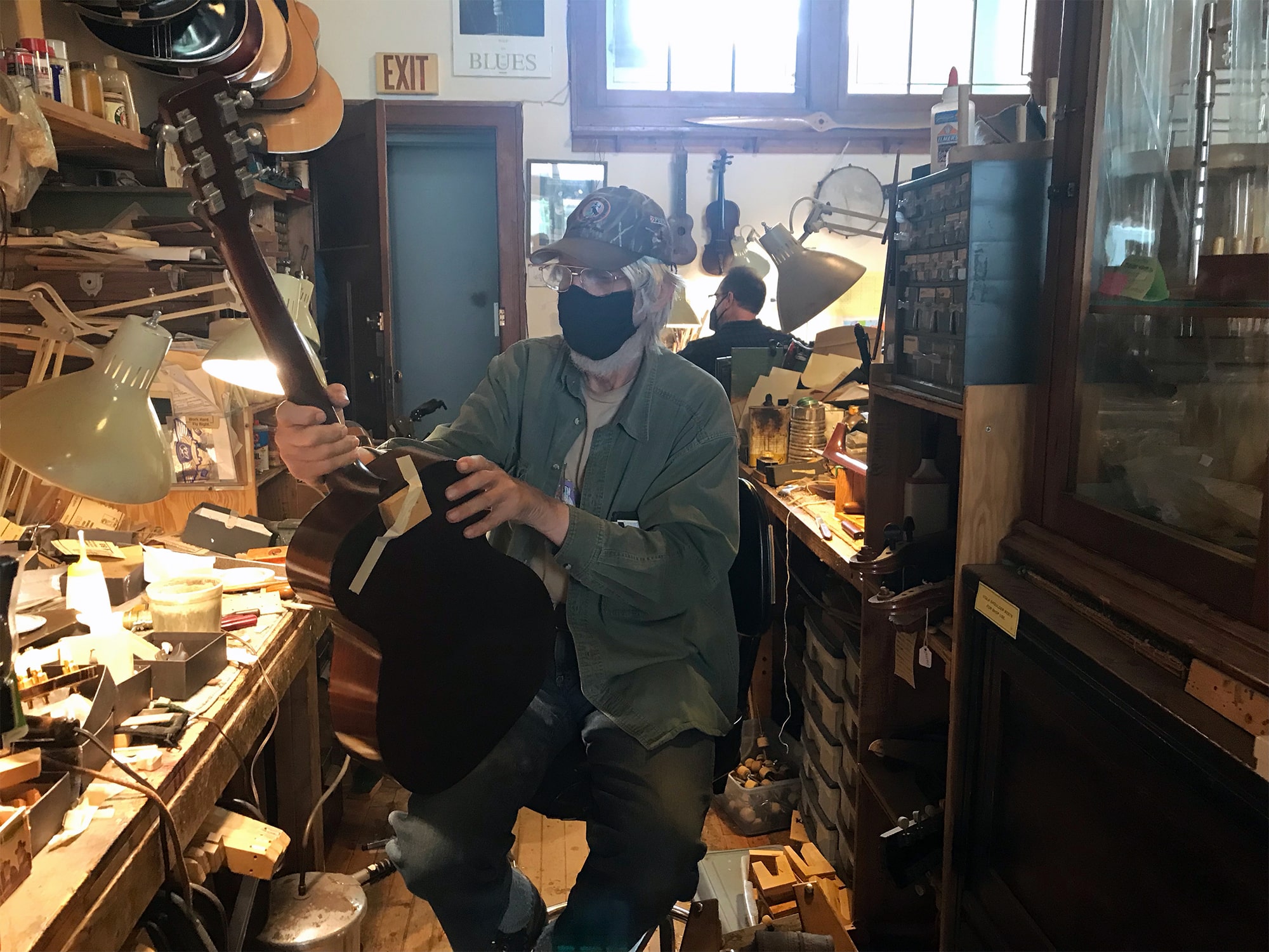 Spruce Tree Music owner Wil Bremer is shown inspecting an acoustic guitar at a crowded workbench at the store. Over his shoulder in the background, employee Doug Meihsner can be seen from behind, working at another workbench in the store. Photo by Andy Moore.