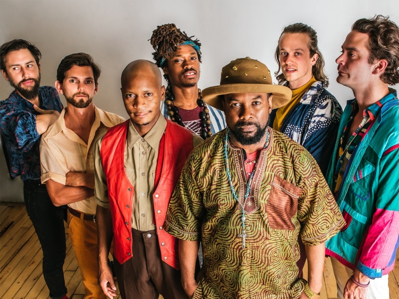 A publicity photo shows seven members of the musical act Kaleta & Super Yamba Band, standing in two rows, wearing bright clothes. Five of the members are facing the camera and two are looking somewhere off to the side.