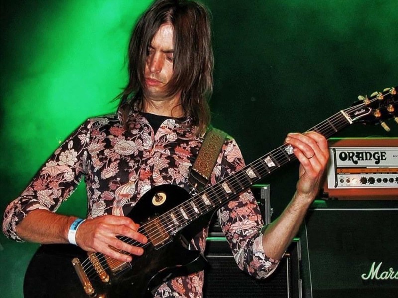 Jason Hartman is pictured playing a 2013 festival in Germany as a member of Jex Thoth. Hartman's playing a black Les Paul and wearing a floral-print black, pink, and white shirt. His hair's long and dark. There's a Marshall amp cab and an Orange head shown to his right. Photograph by Micha.