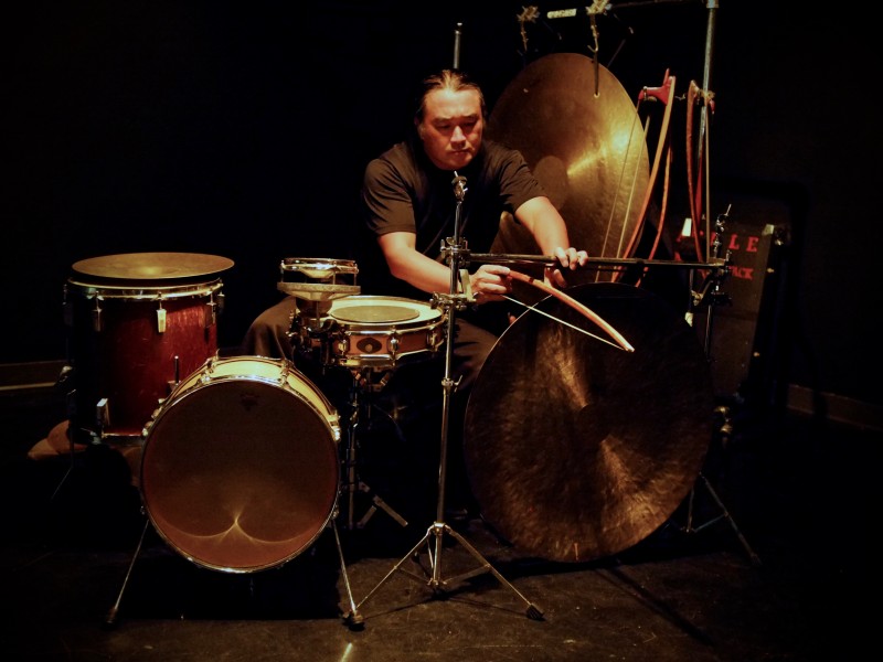 A photo shows percussionist Tatsuya Nakatani using a bow to coax sound from a gong. He is surrounded by a drumkit and another, larger gong hangs from a frame behind him.