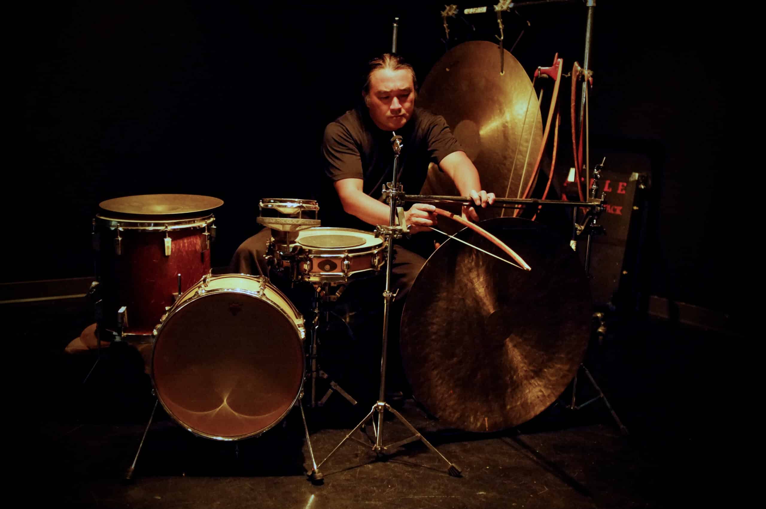 A photo shows percussionist Tatsuya Nakatani using a bow to coax sound from a gong. He is surrounded by a drumkit and another, larger gong hangs from a frame behind him.
