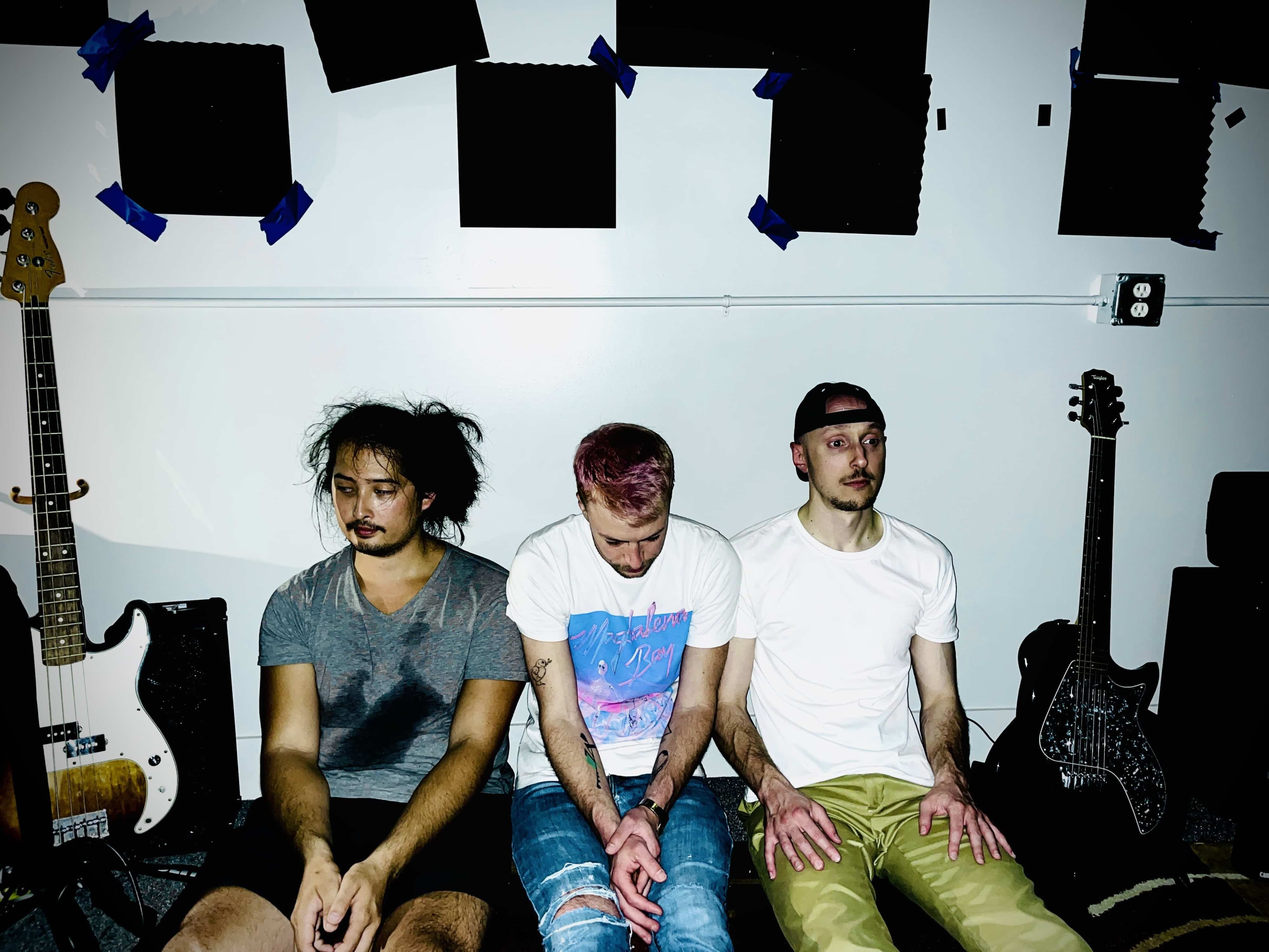 A promotional photo of the band Jonathon Millionaire shows three band members sitting down against the wall of a plastic space, with guitars and sound-treatment panels in the background.
