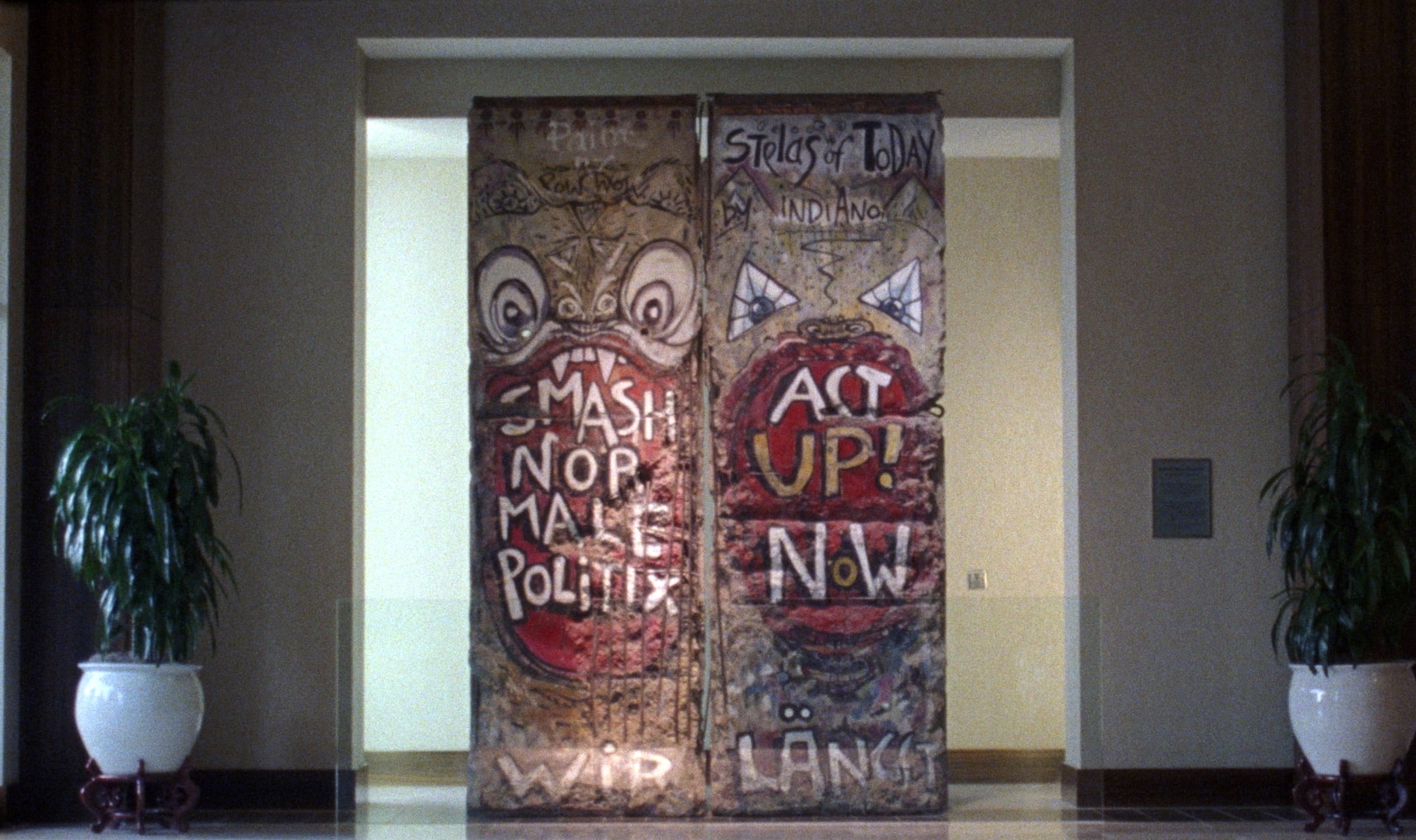A still from the film "The American Sector" shows a section of the Berlin Wall in a room with potted plants on either side.