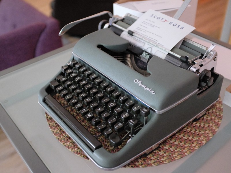Scot Ross' gray-green Olympia typewriter is shown sitting on a desk. A sheet of paper with Ross' letterhead sticks halfway out of the top of the typewriter, with illegible typing on it.
