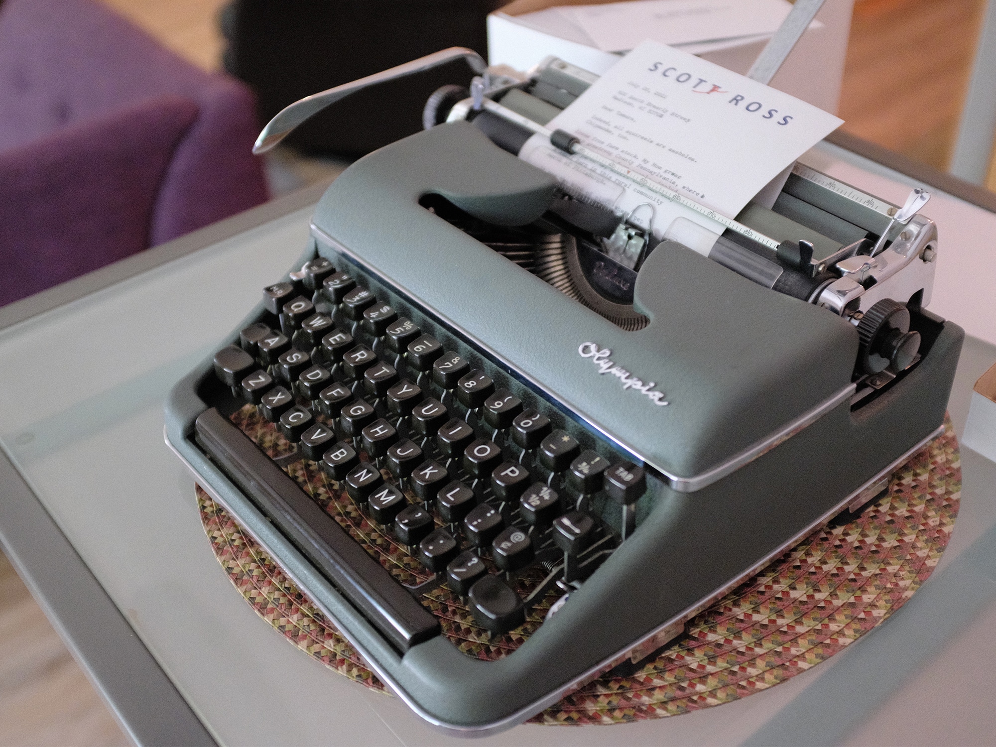 Scot Ross' gray-green Olympia typewriter is shown sitting on a desk. A sheet of paper with Ross' letterhead sticks halfway out of the top of the typewriter, with illegible typing on it.