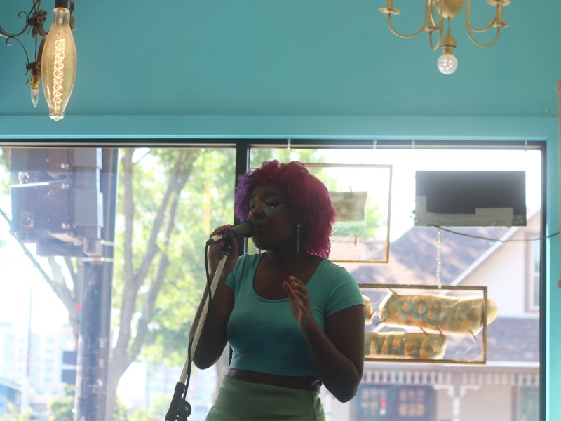 Musician Danielle Crim is shown performing in front of a window at Mother Fool's in Madison. Crim is singing into a microphone, and through the window, trees and buildings on Willy Street are visible.