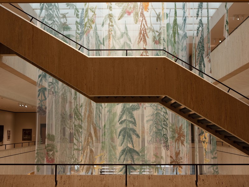 A photo of Amanda McCavour's large scale "Ode To A Prairie" shows the artist's large-scale textile representations of plants and flowers, hanging on translucent sheets of fabric in an atrium at the Chazen Museum of Art. A staircase angles through the foreground of the photo, emphasizing the array of vantage points from which museum visitors can view the work.