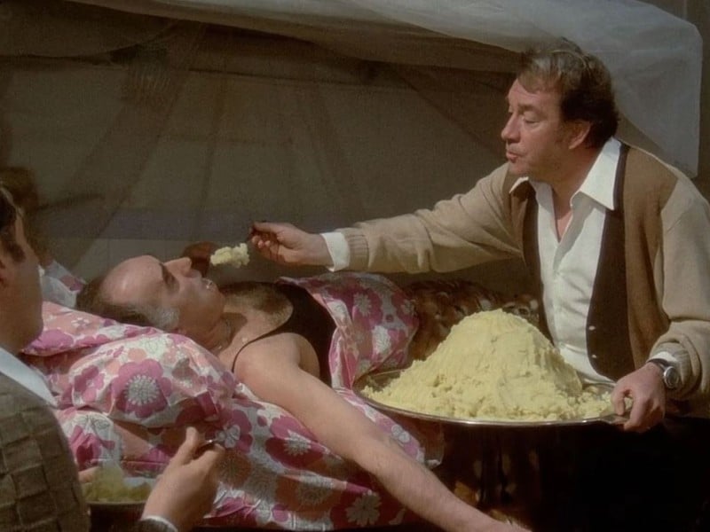Ugo (Ugo Tognazzi) feeds pâté to a bedridden Michel (Michel Piccoli) while Philippe (Philippe Noiret) observes them and eats his own serving of pâté.