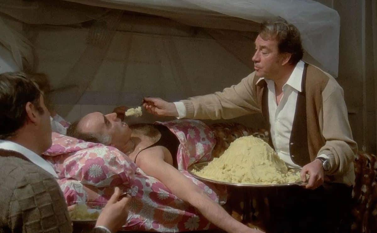 Ugo (Ugo Tognazzi) feeds pâté to a bedridden Michel (Michel Piccoli) while Philippe (Philippe Noiret) observes them and eats his own serving of pâté.