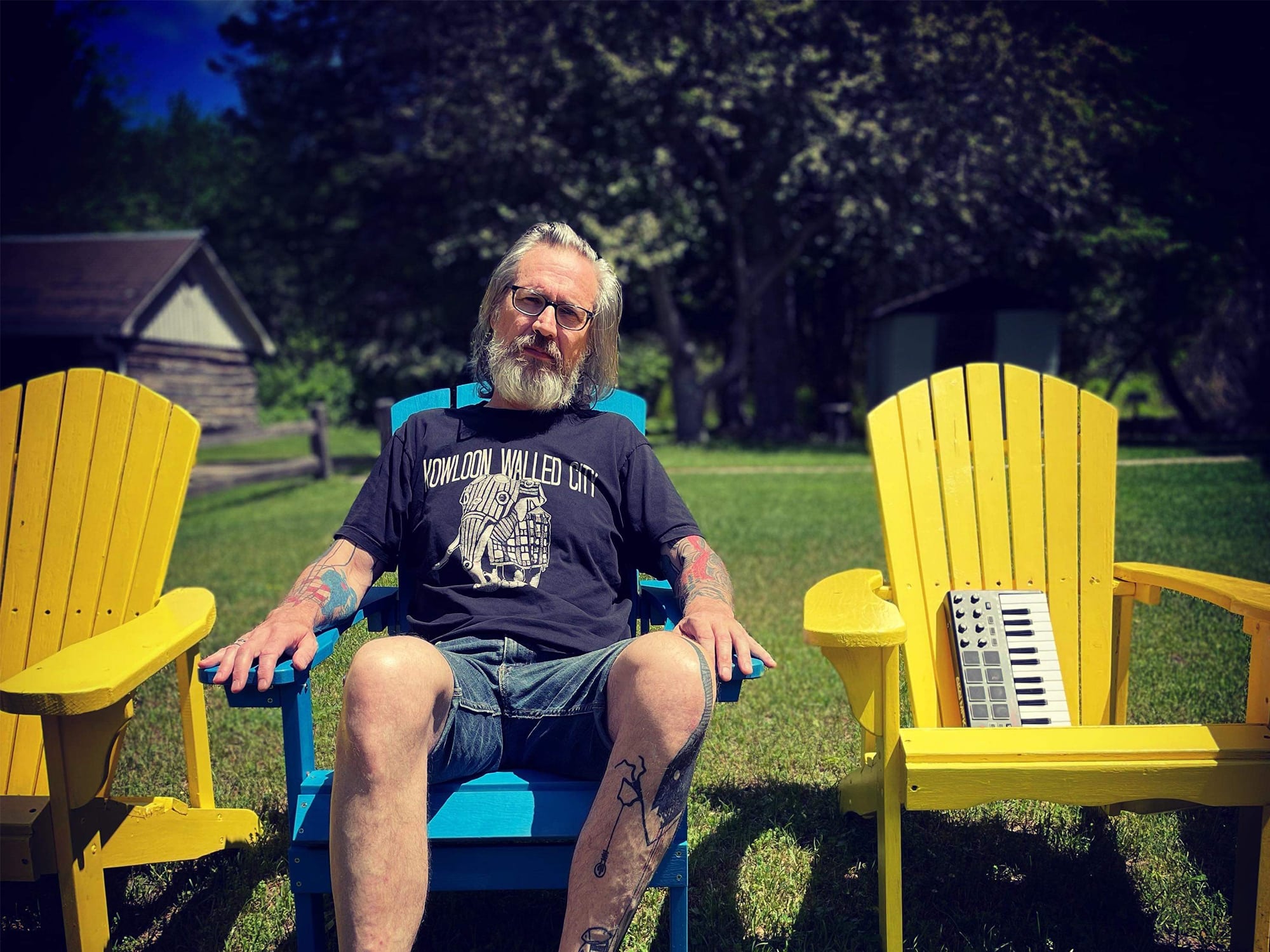 Musician Russell Emerson Hall is shown sitting on a lawn chair with a yard and trees in the background. Next to him is another chair, with a MIDI controller keyboard sitting in it.