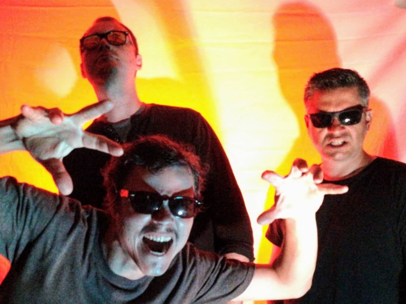 A publicity photo of the band Sinking Suns shows the band's three members posing in sunglasses against a bright orange background.