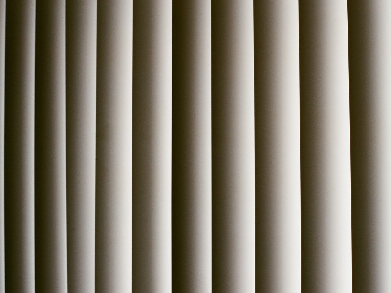 An analog photograph of vertical blinds. The blinds take up all the space of the image. While light comes through them, you cannot see beyond them.