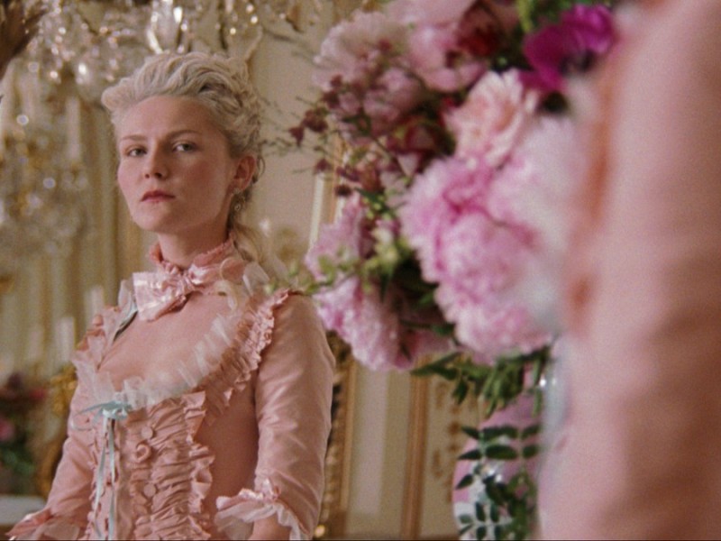 In a frilly, peach-colored, Georgian era dress, Marie Antoinette (Kirsten Dunst) gives an aggrieved look at herself in the mirror. A vase of pink flowers sits to her right on the dresser in the middle ground of the shot.