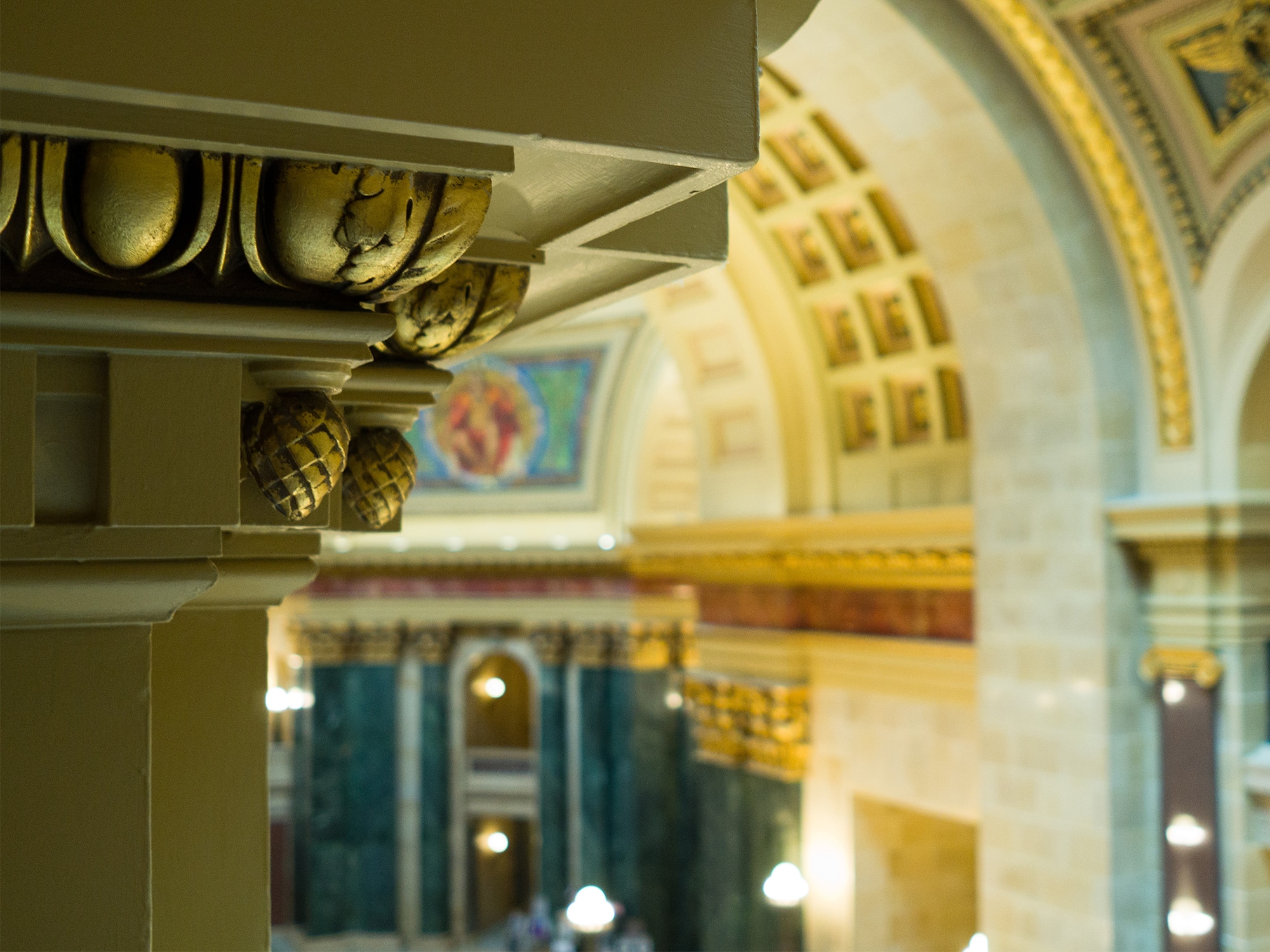 A photo of the inside of the Wisconsin State Capitol rotunda shows the top of a column-like formation on an upper floor in the left foreground. In the background, out of focus, the building’s vaulted ceiling and an archway decorated with a mosaic are visible.