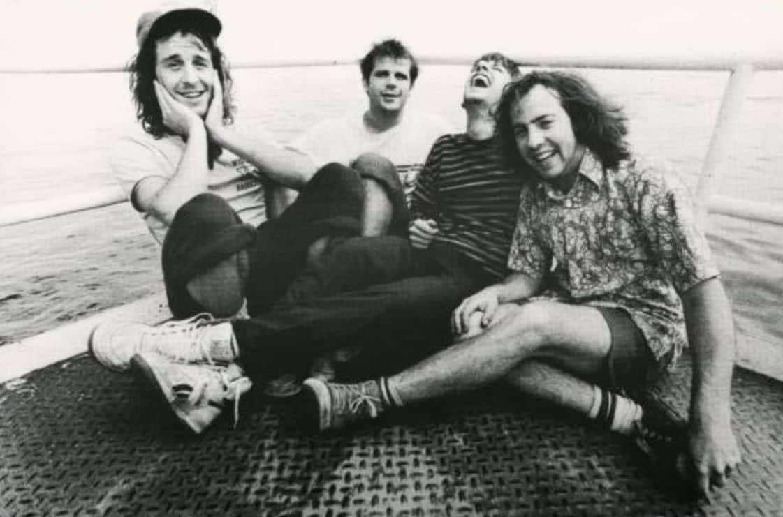 An old black-and-white publicity photo shows the members of the band Poopshovel sitting on a dock and smiling with a body of water in the background.