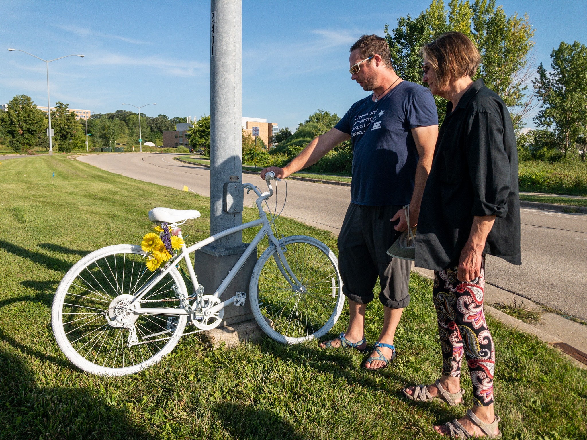 A bicycle painted white with yellow flowers tied to the back of the seat post leans against a metal pole in a grassy median between two roads. Will Schira wears a blue shirt, pants, and sandals and is grasping one handlebar. Charlotte Cummings stands next to him, looking at the bike, and wears a long black shirt and paisley leggings.