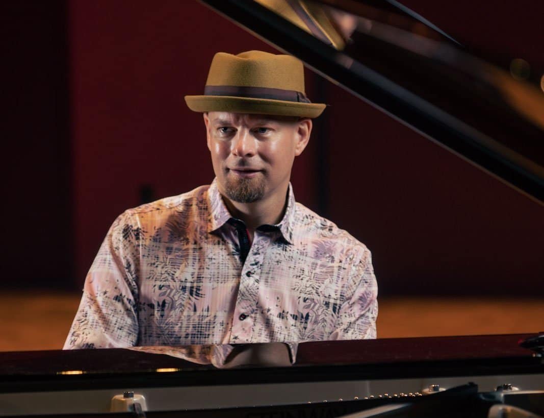 A publicity photo shows Johannes Wallmann playing a grand piano. His head and shoulders are framed by the piano's open lid.