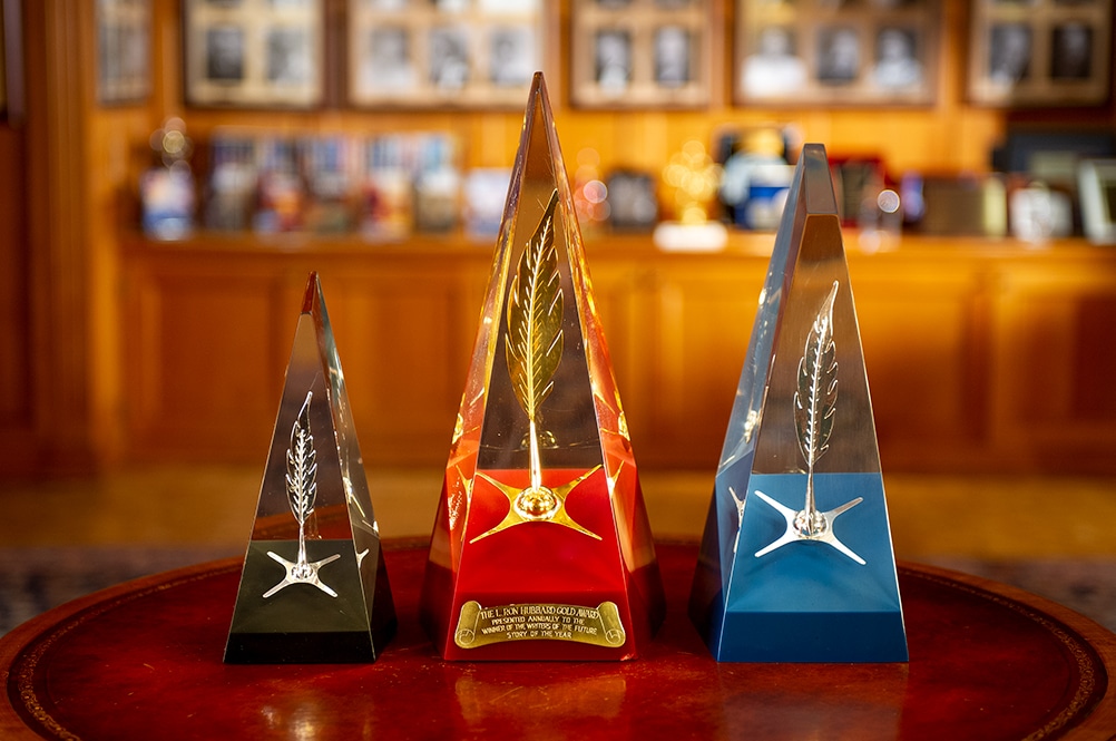 A photo shows three award trophies from the Writers Of The Future contest. The trophies are large, triangular objects consisting of a glass pyramid atop a solid-colored base. Embedded in the glass portion of each is a feather.