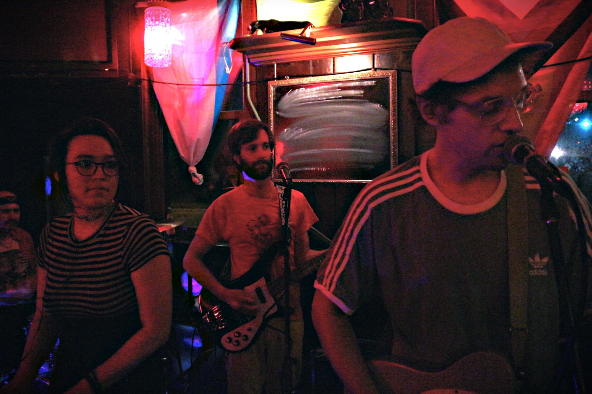 Photo: Graham Hunt performs with a full backing band at Mickey's Tavern in June 2022 to celebrate the release of his latest album. Left to right: Sam Reitman (drums), Emili Earhart (keys), Stephen Strupp (bass), and Graham Hunt (guitar/vocals). Not pictured, to Hunt's left, are Shannon Connor (guitar) and Isaac deBroux-Slone (acoustic guitar, percussion). The photo has an orange-ish hue and a slight grain effect.