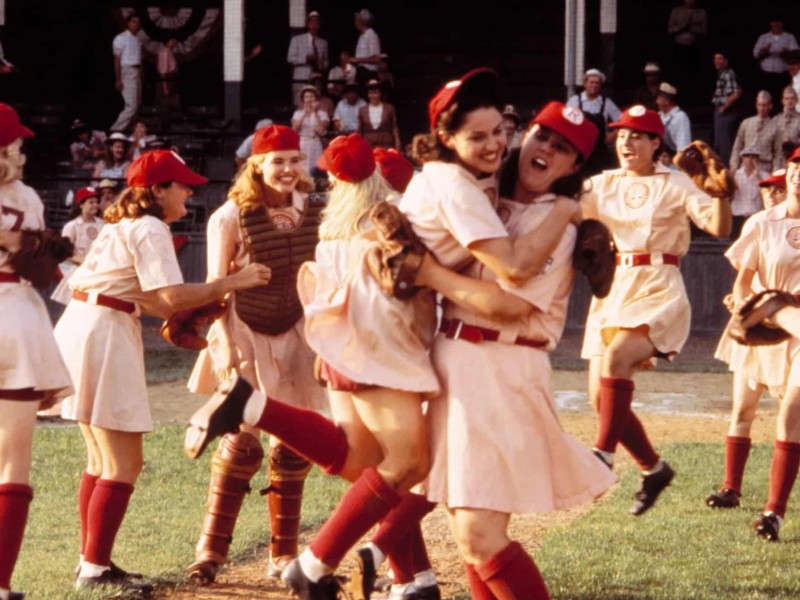 The Rockford Peaches celebrate a nail-biting win on the field. They wear their gloves and other equipment with their pink, short-skirt uniforms and baseball caps. Mae (Madonna) jumps into the arms of her best friend Dorris (Rosie O'Donnell) while Dottie (Geena Davis) laughs and cheers with the rest of the team.