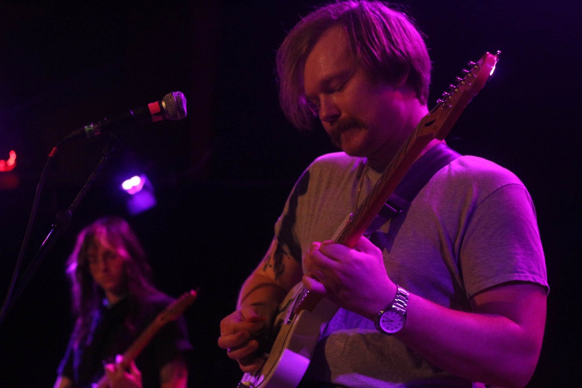 Proud Parents' Tyler Fassnacht is shown in focus, to the right of the image. He's wearing a gray shirt and his eyes are closed as he plays. In the background, C Nelson-Lifson can be seen in soft focus. Nelson-Lifson's eyes are also closed as they play.