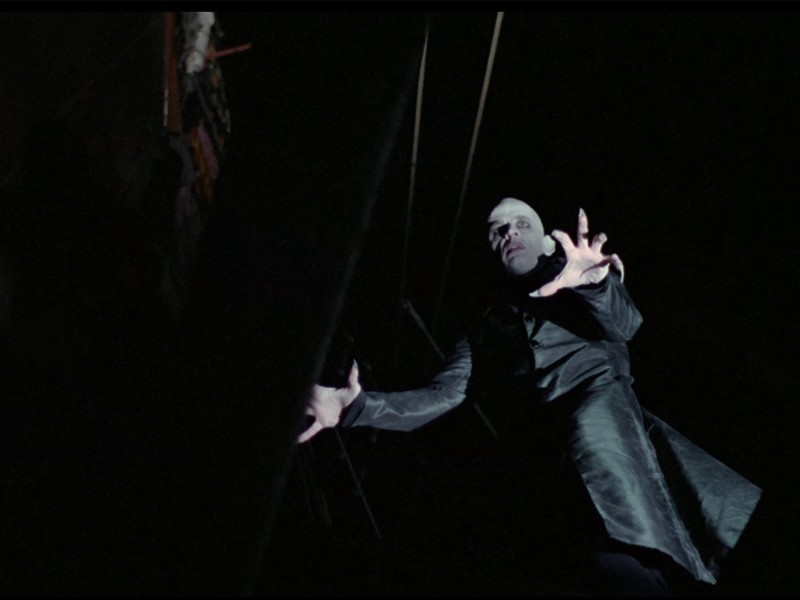 In a still from Nosferatu: Phantom Of The Night, Dracula, played by Klaus Kinski, is shown from below with a dramatically tilted camera angle. Dracula, deathly pale and clad in a flowing black garment, has extended his fingers, which have long pointy nails.