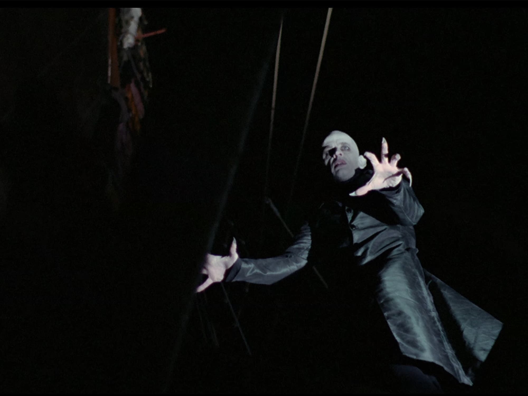 In a still from Nosferatu: Phantom Of The Night, Dracula, played by Klaus Kinski, is shown from below with a dramatically tilted camera angle. Dracula, deathly pale and clad in a flowing black garment, has extended his fingers, which have long pointy nails.