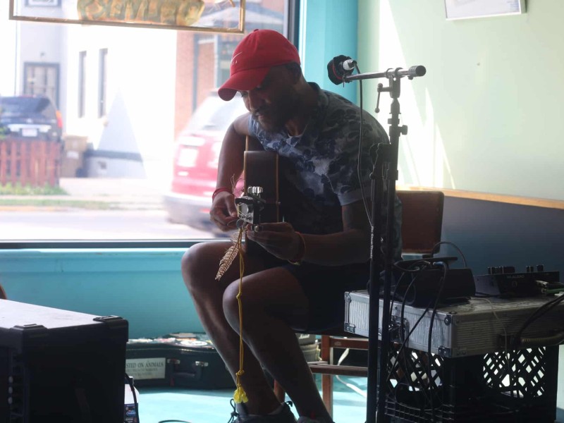 Def Sonic is shown tuning his acoustic guitar at Mother Fool's during Make Music Madison. He's wearing a red Nike baseball cap and a gray shirt with a blue floral print. Next to him is a makeshift table, created by crates and an iron briefcase, holding up pieces of additional audio equipment.