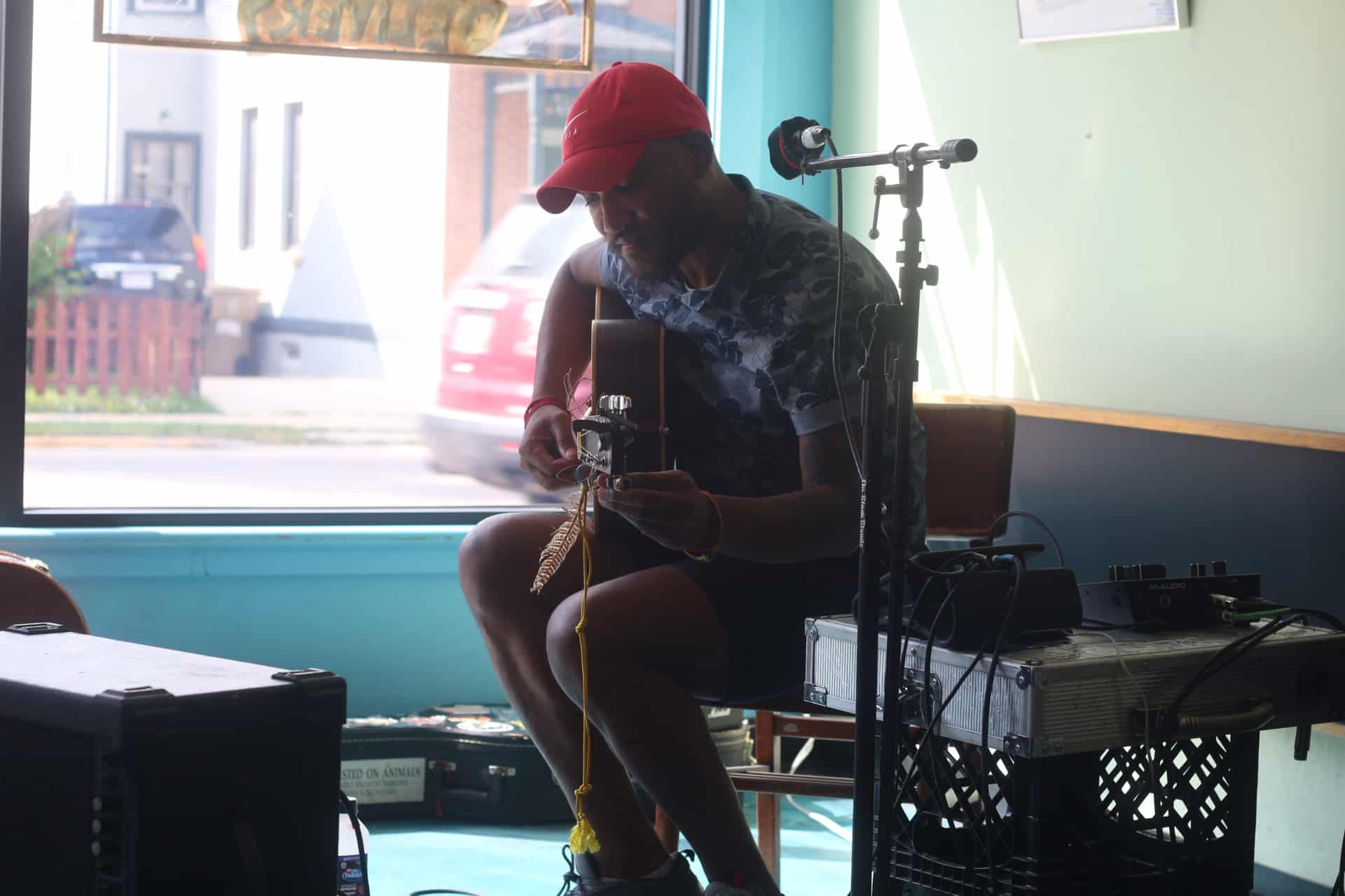 Def Sonic is shown tuning his acoustic guitar at Mother Fool's during Make Music Madison. He's wearing a red Nike baseball cap and a gray shirt with a blue floral print. Next to him is a makeshift table, created by crates and an iron briefcase, holding up pieces of additional audio equipment.