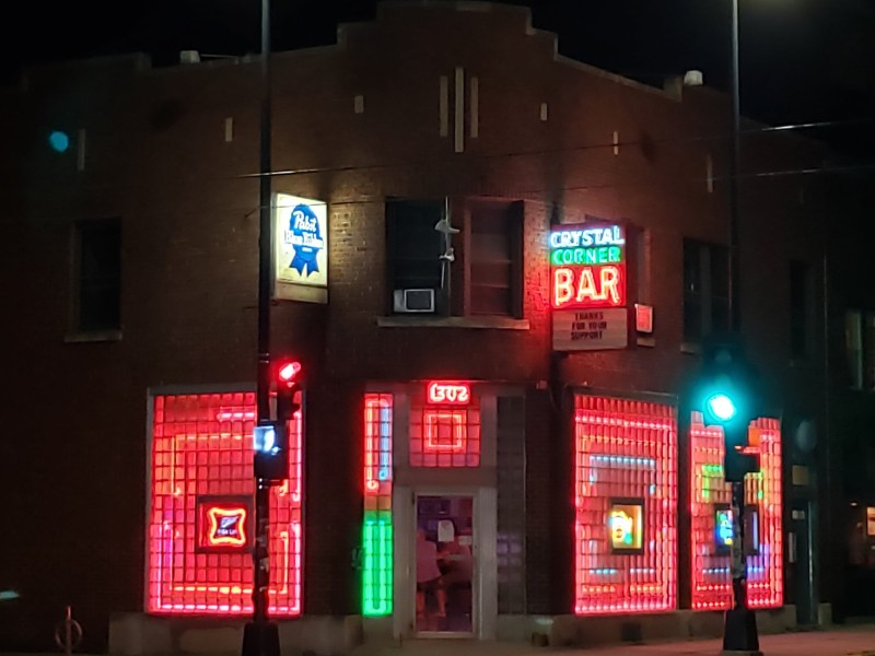 The entrance of the Crystal Corner Bar is shown from a kitty-corner angle, emphasizing the glass-brick exterior backlit with red lights.