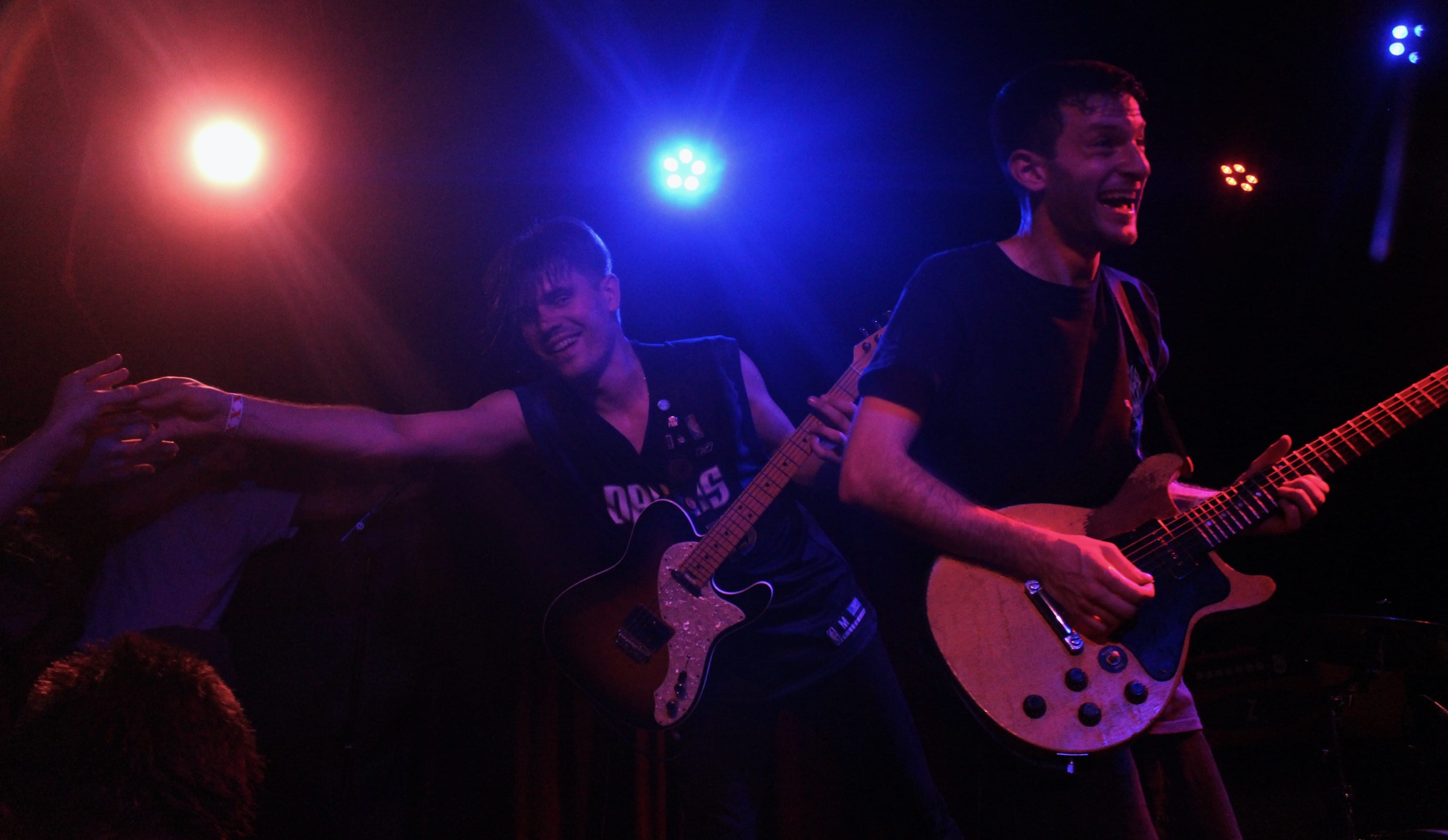 Two members of the band PUP are shown, playing guitar, and smiling. PUP's lead guitarist, Steve Sladkowski, is in the background, leaning over to high-five a crowd member.