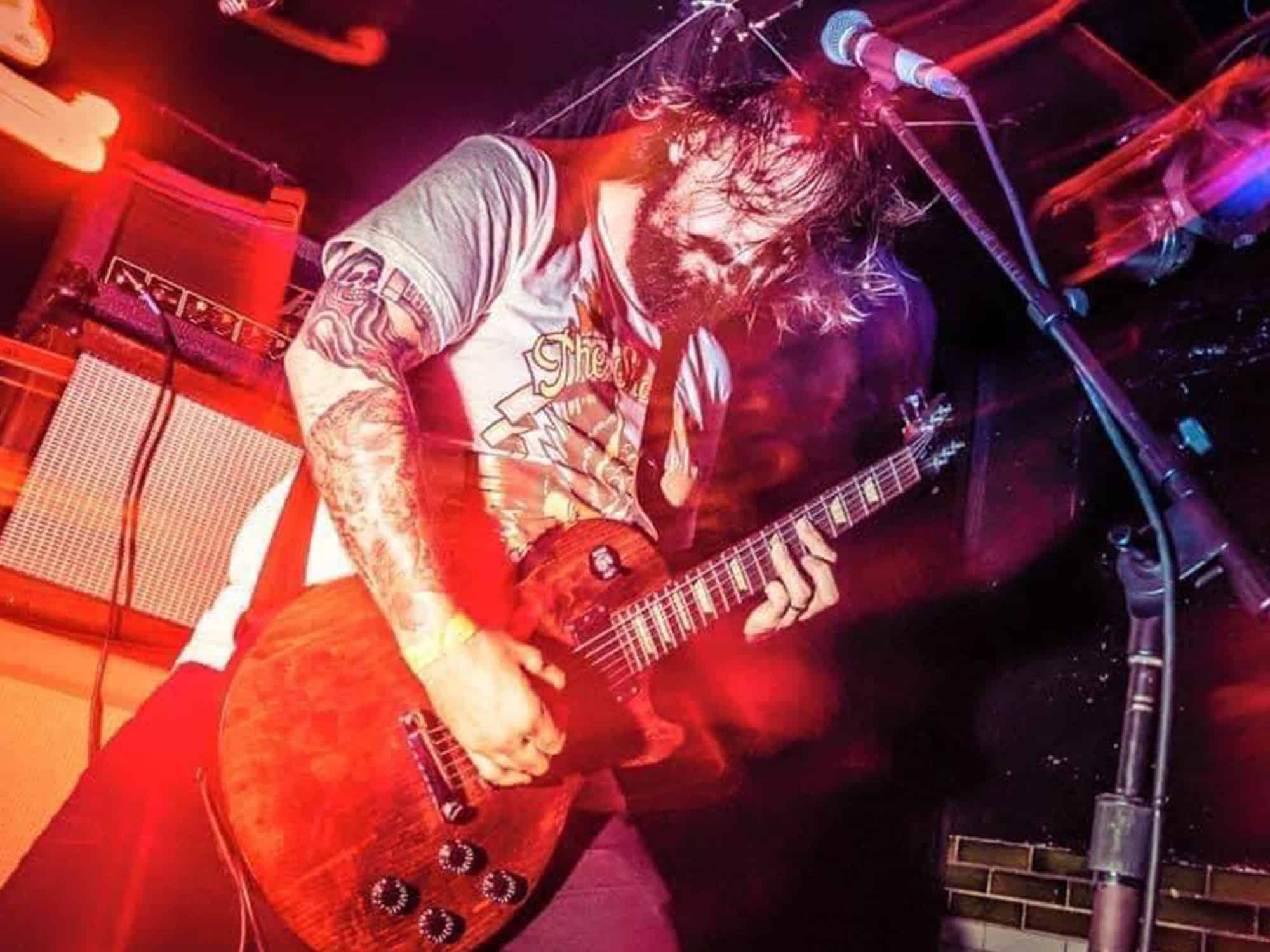 A photo shows Alex Linden playing guitar during a show with this band Bereft, his eyes closed and his hair swinging about his head as he stands in a lunge-like posture.