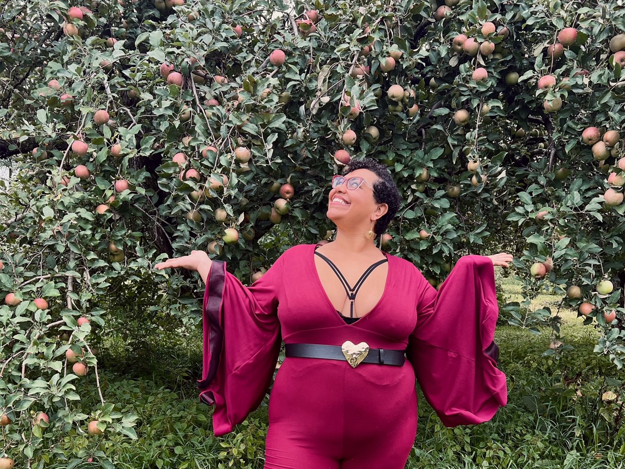 The author is shown posing in an apple orchard and wearing a red outfit with long billowy sleeves.