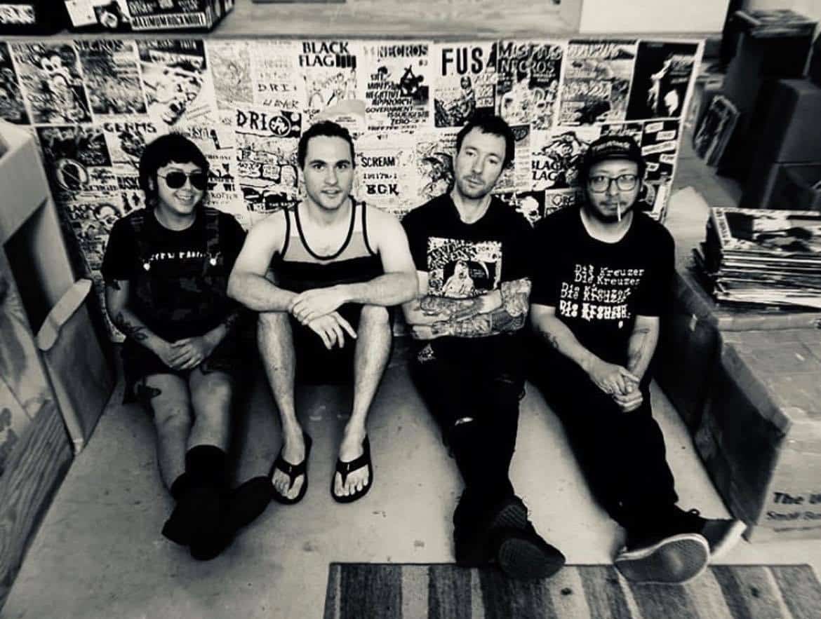 The four members of No Question are shown posing for a photo, seated on a floor with their backs against a wall covered with punk show flyers.