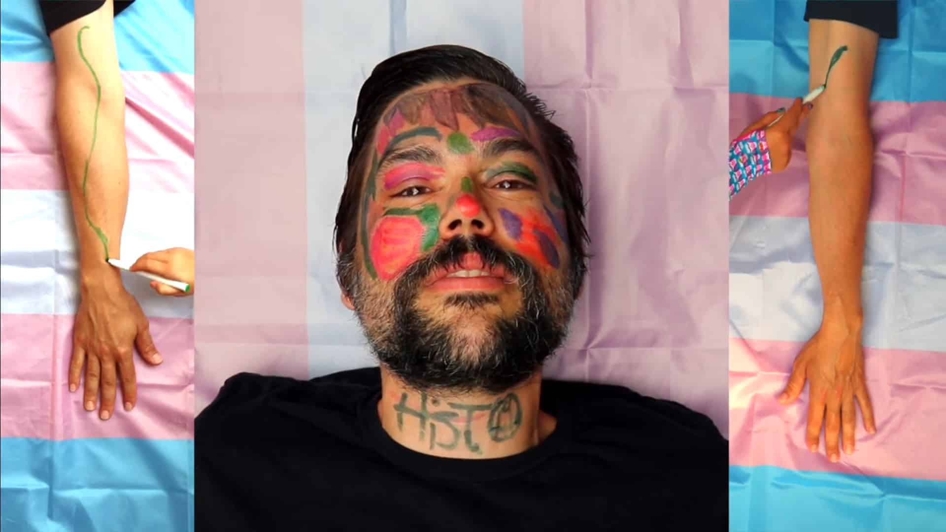 Histo's Donald Curtis' face is shown in the center of the image. The image is split into three panels. The side panels show Curtis' arms as they are painted on by children. The middle panel shows Curtis' face, which is also heavily painted. "Histo" is written across his neck. Underneath him, in all panels, the transgender flag is visible.